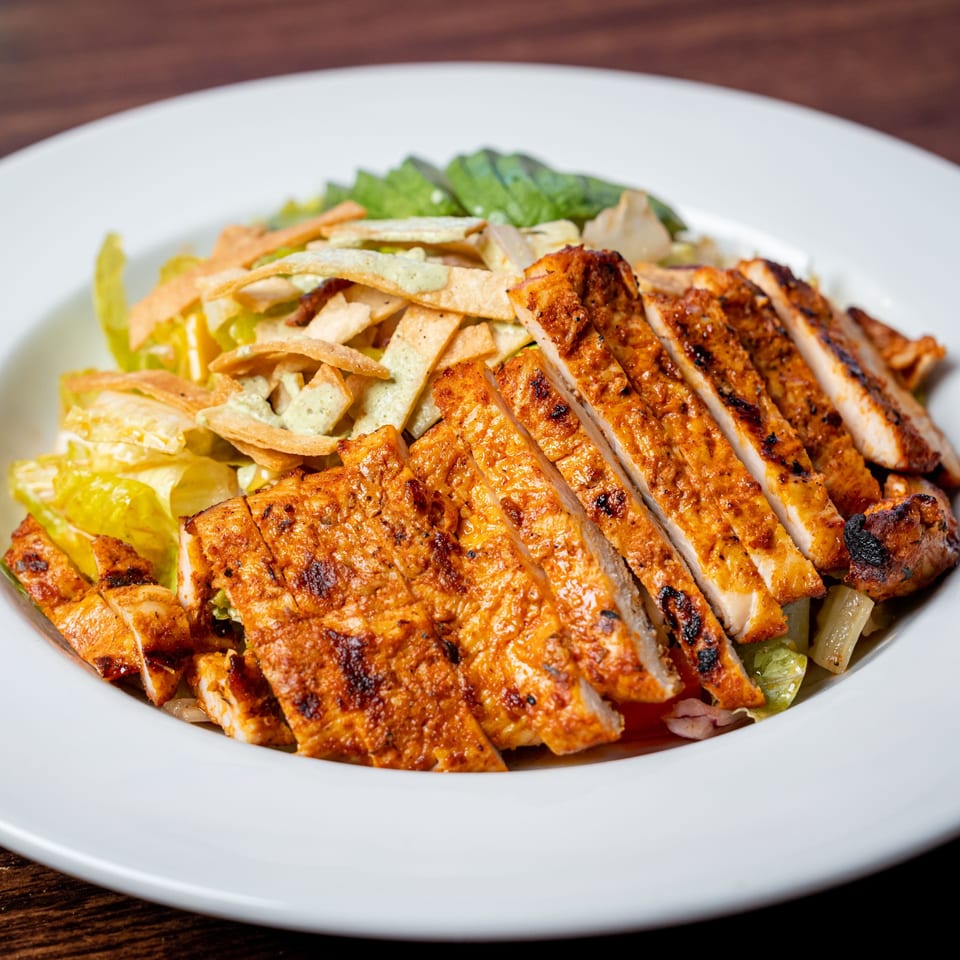 Grilled chicken salad with lettuce, crispy tortilla strips, and a creamy dressing on a white plate.