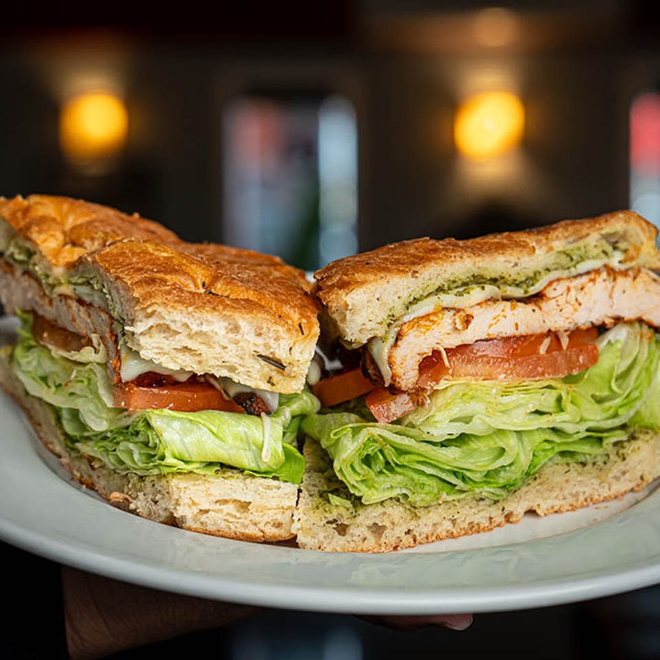 A large chicken sandwich, cut in half, with lettuce, tomato, and pesto on focaccia bread.