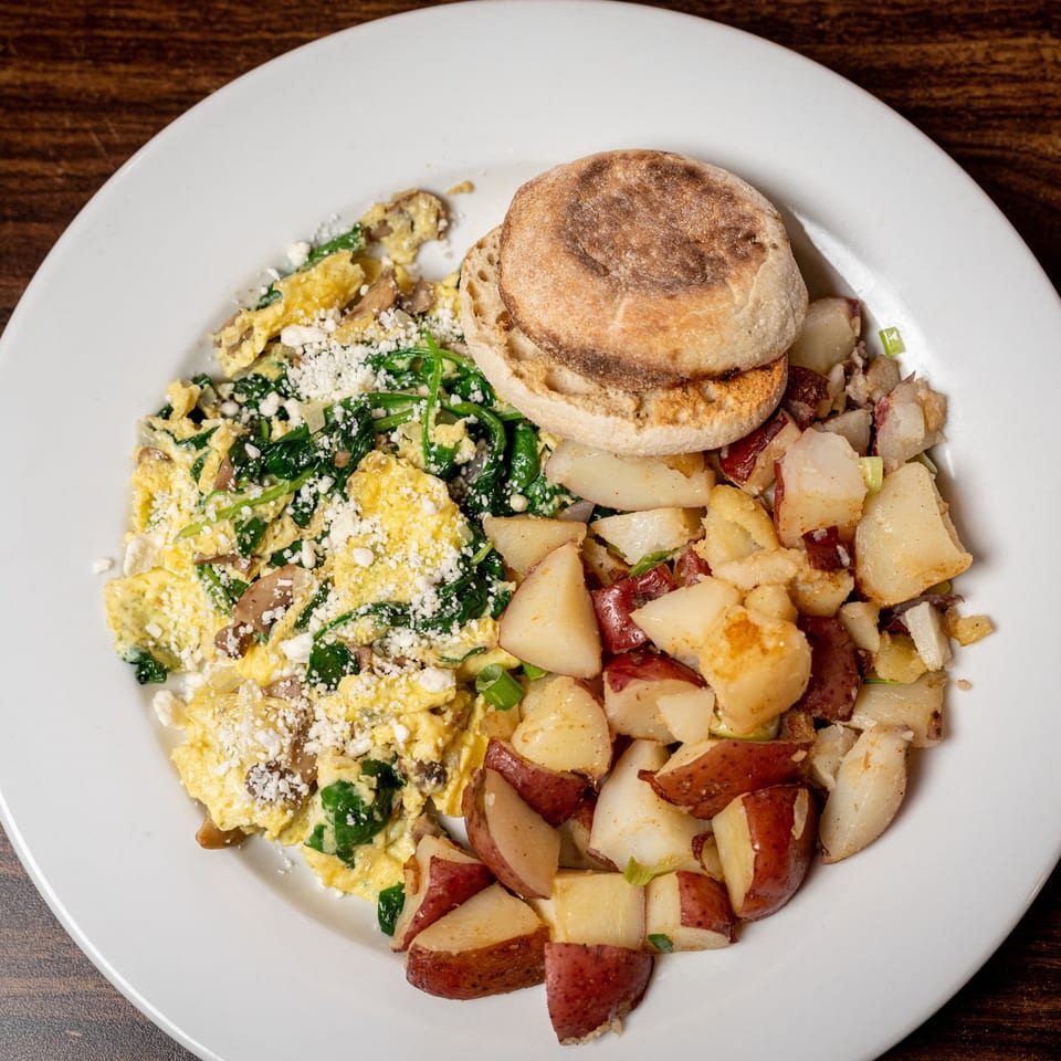 Scrambled eggs with spinach and mushrooms, home fries, and an English muffin on a white plate.